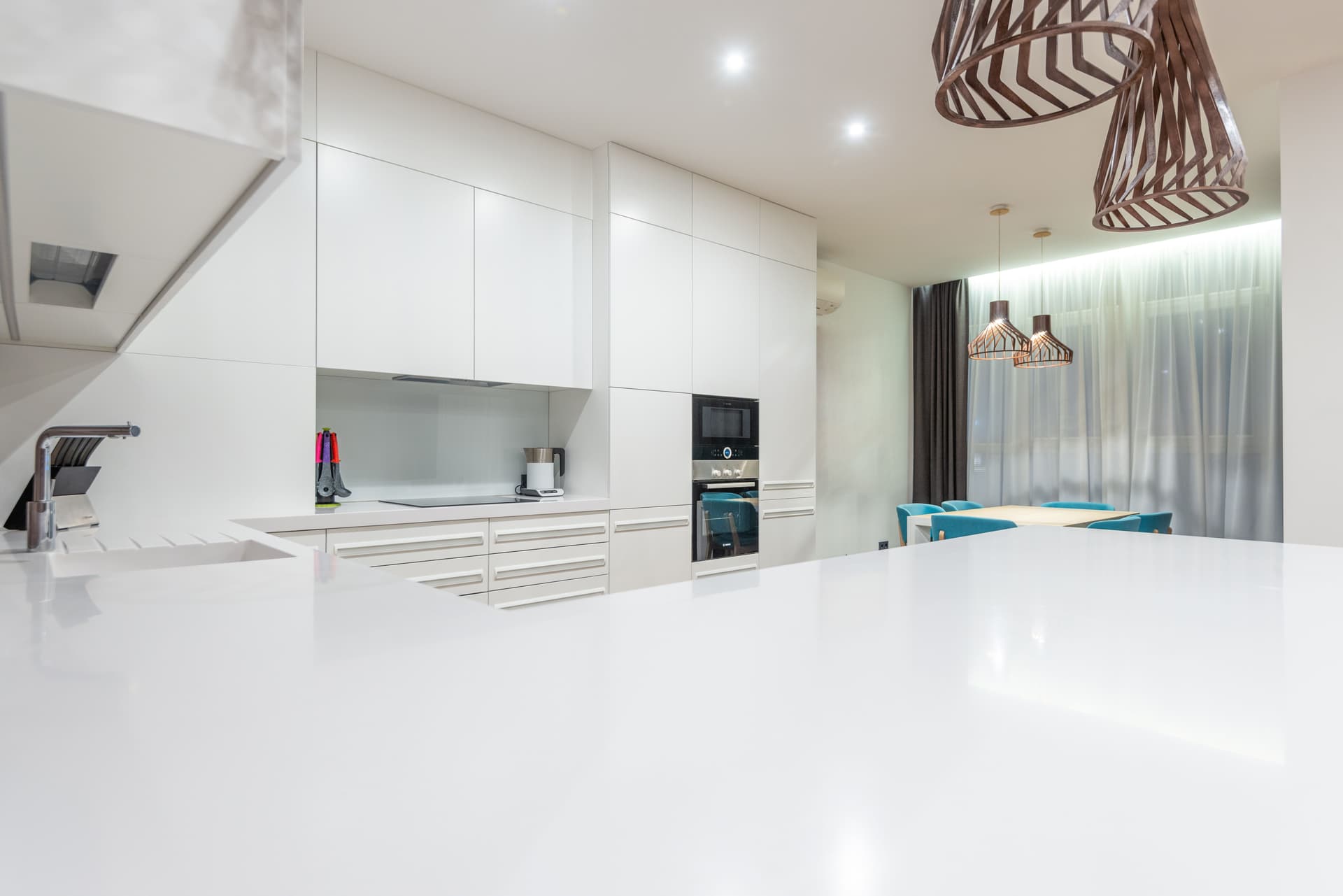 An empty light kitchen counter running along white cupboards, with a window and a dining table further back in the room.